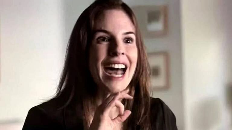 Mulher de cabelos castanhos reage com entusiasmo e boca aberta enquanto fala em ambiente interno iluminado em cena de novela ou série