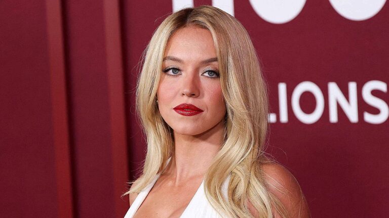 Sydney Sweeney posa diante de painel vermelho durante evento de estreia com vestido branco e batom vermelho