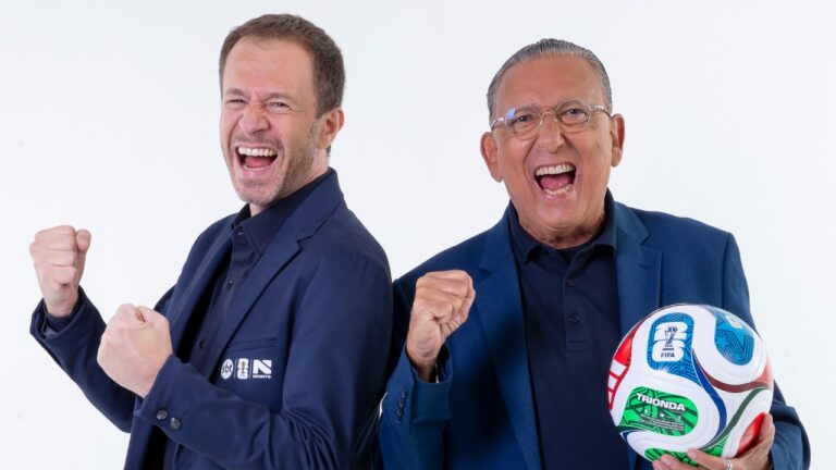 Tiago Leifert e Galvão Bueno de blazer azul comemoram com punhos cerrados enquanto posam com bola de futebol em foto promocional do SBT
