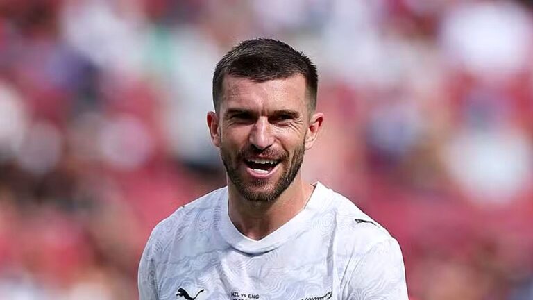 Jogador de futebol de uniforme branco sorri durante partida em estádio com arquibancadas desfocadas ao fundo e expressão de comemoração