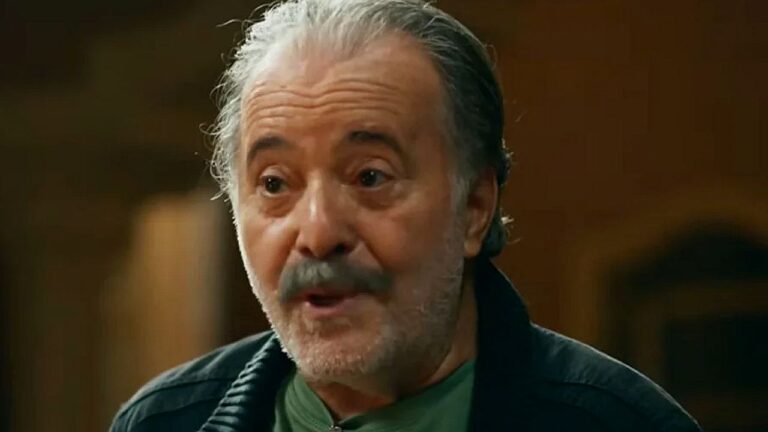 Homem grisalho de barba e bigode fala com expressão intensa durante conversa em ambiente interno iluminado em cena de novela