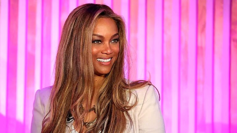 Tyra Banks sorri durante participação em evento com iluminação rosa ao fundo e blazer claro
