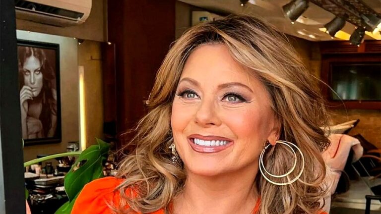 Vera Fischer sorri durante entrevista usando blusa laranja e brincos de argola em camarim de estúdio