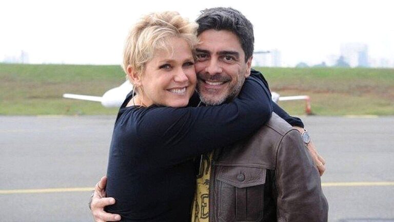 Xuxa Meneghel abraça Junno Andrade e sorri para a câmera em área de aeroporto com avião desfocado ao fundo