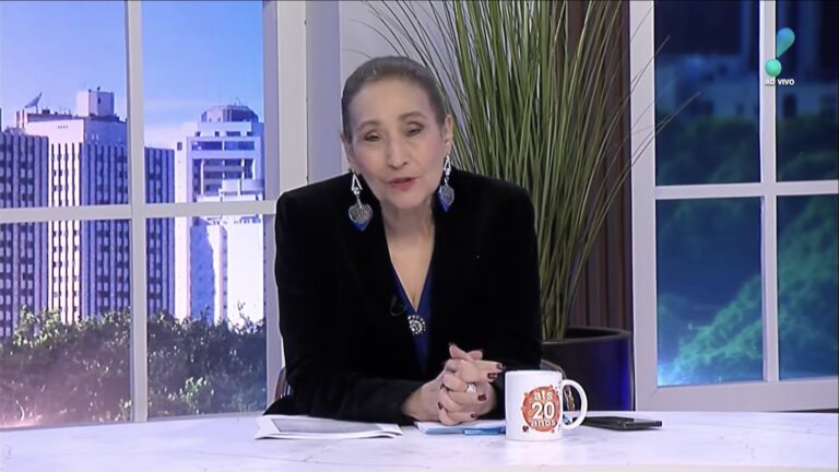 Sonia Abrão, de blazer preto, na bancada do A Tarde é Sua, da RedeTV!, com caneca, papéis e janela com prédios ao fundo