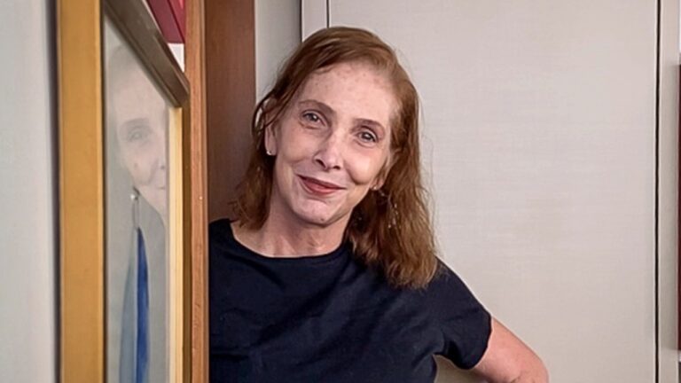 Adriana Falcão sorri apoiada no batente da porta, veste blusa preta e aparece ao lado de quadro com seu reflexo visível