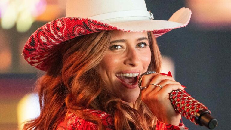 Agrado (Isadora Cruz) de chapéu branco canta com microfone em mãos em cena da novela Coração Acelerado