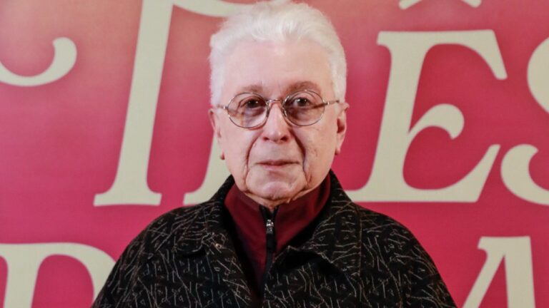 Aguinaldo Silva de óculos e cabelo branco posa diante de painel vermelho com letras claras ao fundo