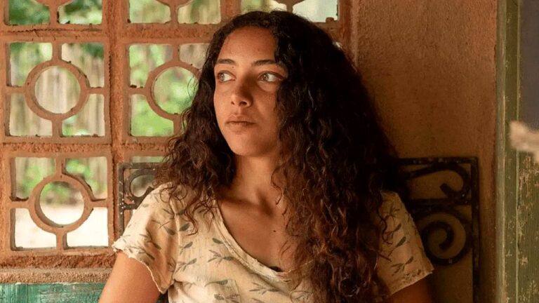 Alice Carvalho com expressão apreensiva encostada na parede em cena de novela