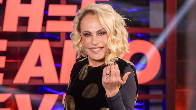 Ana Maria Braga sorri e faz gesto com a mão em estúdio usando vestido preto com círculos dourados e fundo iluminado