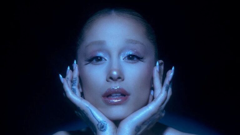 Ariana Grande encara a câmera com maquiagem brilhante e mãos no rosto em imagem promocional da cantora