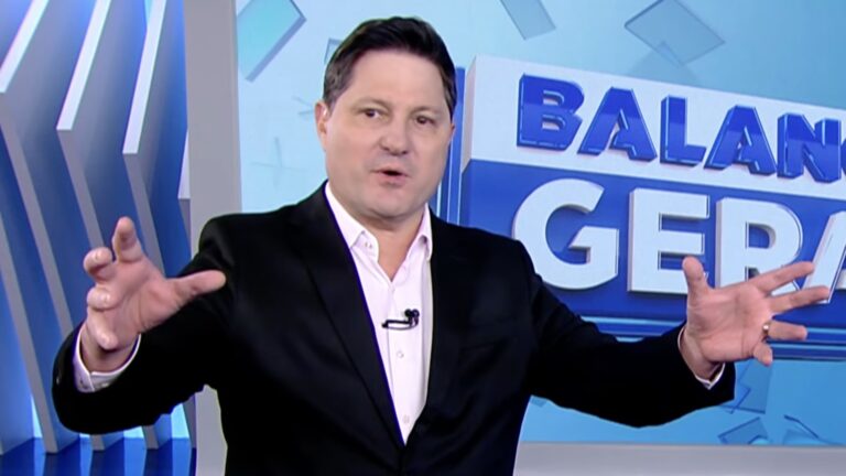 Eleandro Passaia, de terno preto, gesticula no estúdio do Balanço Geral com logotipo do programa ao fundo