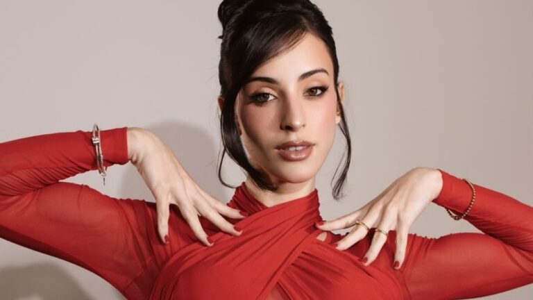 Bella Longuinho posa com vestido vermelho de mangas transparentes e mãos junto ao rosto diante de fundo neutro