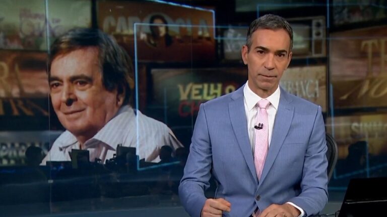 Cesar Tralli de terno azul apresenta telejornal com imagem de Benedito Ruy Barbosa em telão ao fundo no estúdio