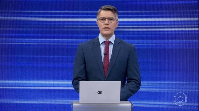 Bruno Tavares, de terno azul e gravata vermelha, atrás de notebook no estúdio do Jornal da Globo