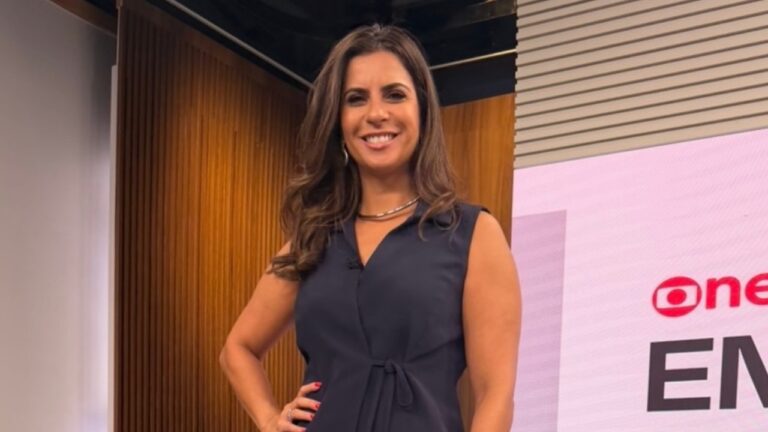 Camila Bomfim sorri de vestido azul-marinho em estúdio da GloboNews diante de telão com logotipo de jornal
