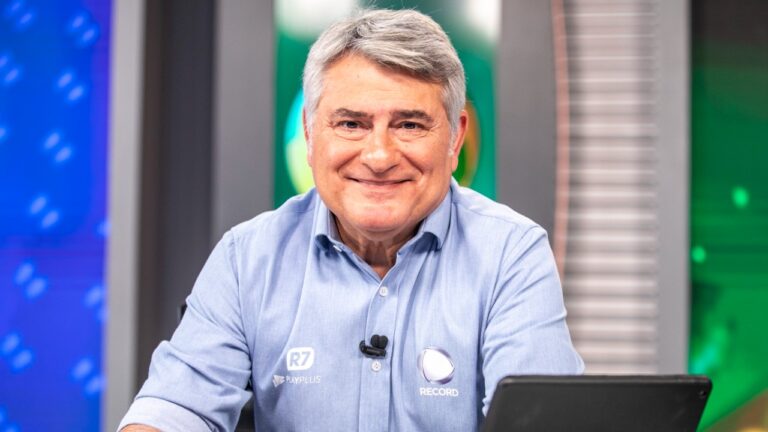 Cléber Machado sorri de camisa azul sentado à bancada com tablet e microfone de lapela em estúdio da Record