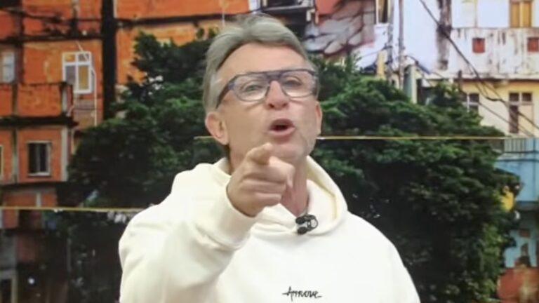 Craque Neto, de moletom branco, aponta para a câmera e grita no estúdio do Os Donos da Bola