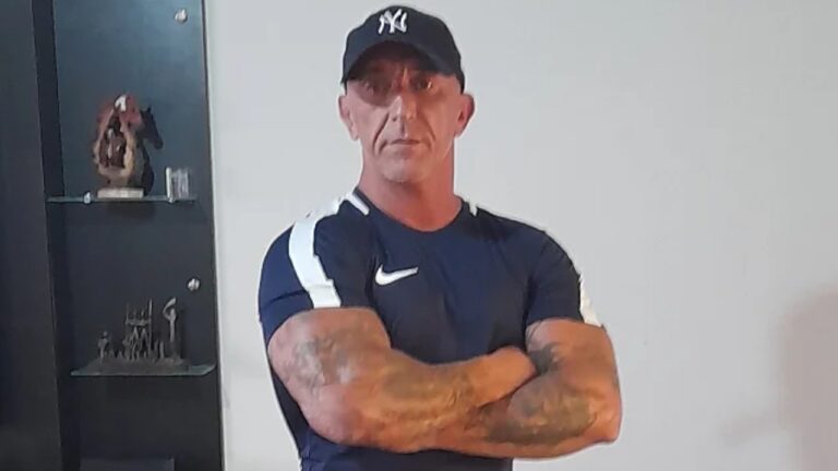 Cristian Cravinhos de boné preto e camiseta esportiva posa com braços cruzados em ambiente interno diante de parede clara