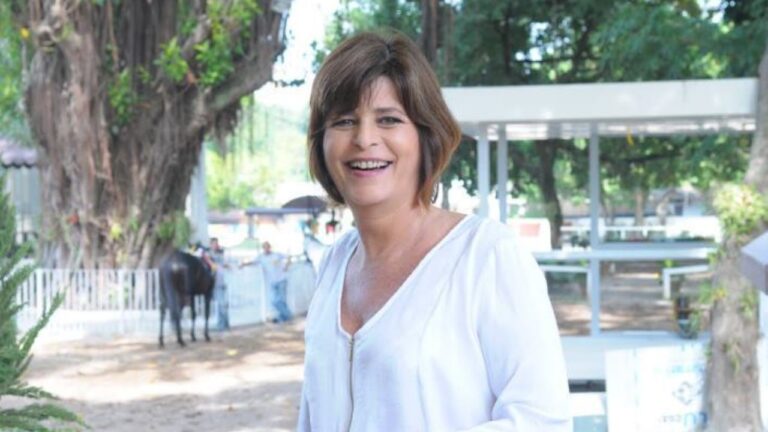 Cristianne Fridman sorri em área externa usando blusa branca com árvores e estrutura clara ao fundo