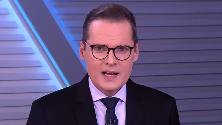 Daniel Caniato de terno escuro e gravata fala diante da câmera em estúdio de telejornal com painel cinza ao fundo