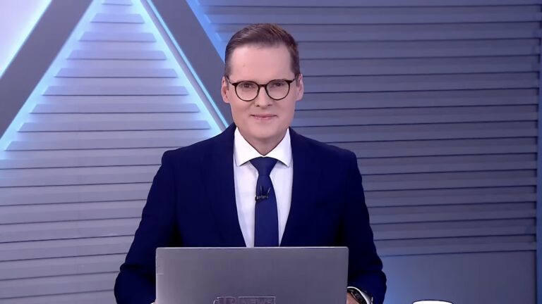 Daniel Caniato, de terno azul atrás de notebook, no estúdio de Os Pingos nos Is, na Jovem Pan