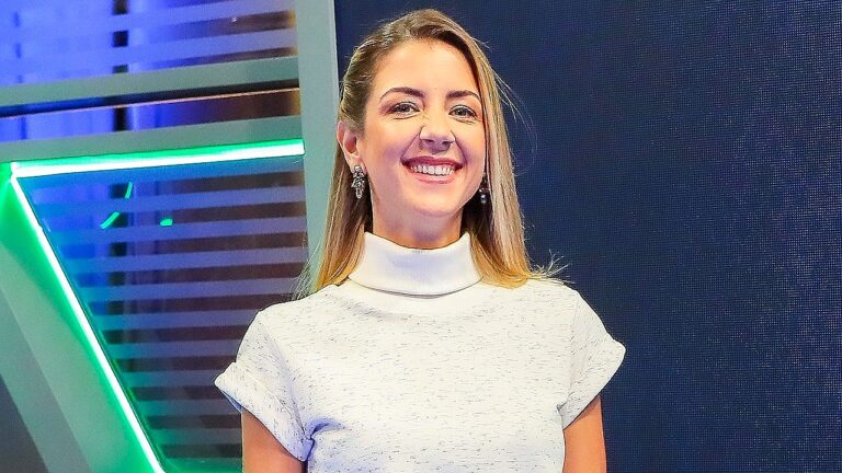 Domitila Becker sorri em estúdio de TV usando blusa clara de gola alta durante apresentação esportiva