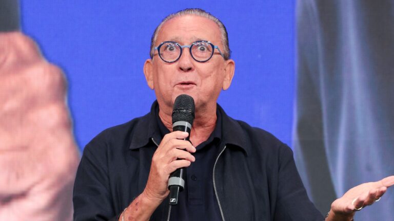Galvão Bueno de óculos redondos segura microfone e gesticula em palco durante evento