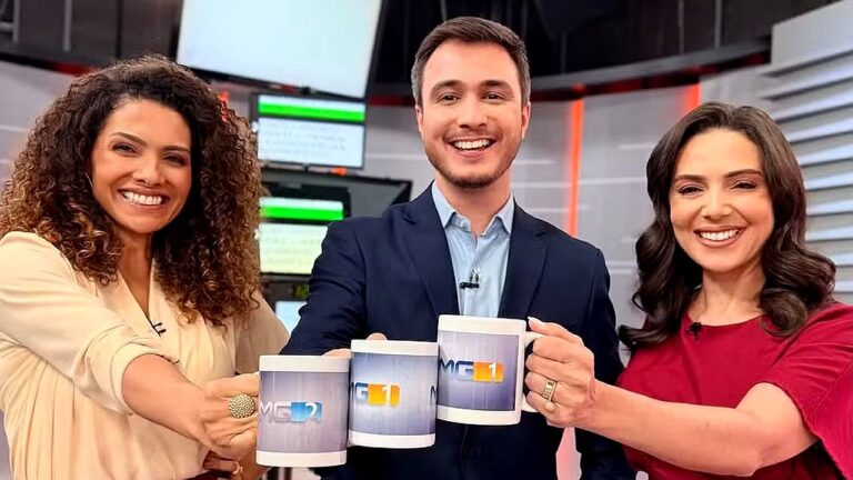 Três apresentadores da Globo sorriem no estúdio de telejornal enquanto erguem canecas personalizadas dos programas MG1 e MG2