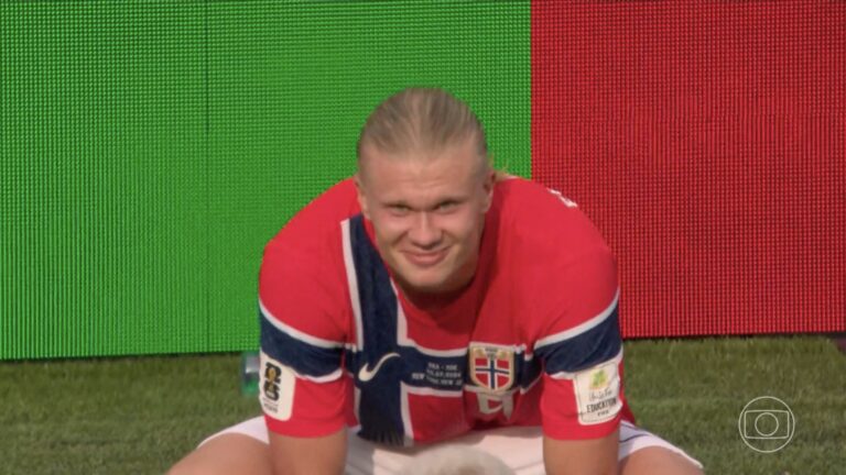 Erling Haaland, agachado e sorrindo, vestindo camisa da Noruega em campo durante Brasil x Noruega