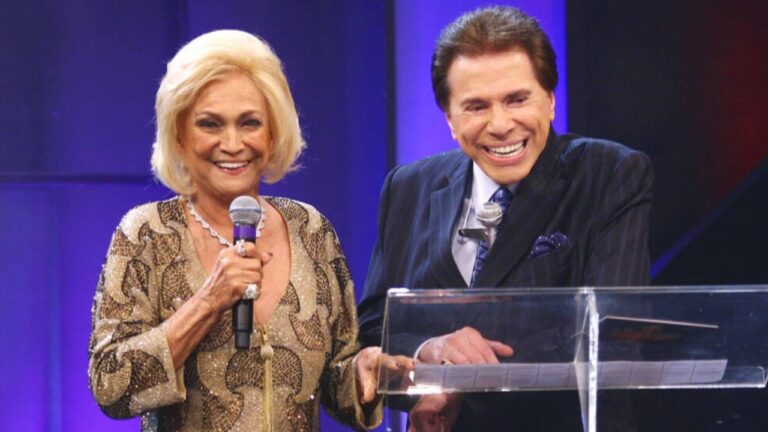 Hebe Camargo de vestido dourado segura microfone ao lado de Silvio Santos em palco com fundo azul