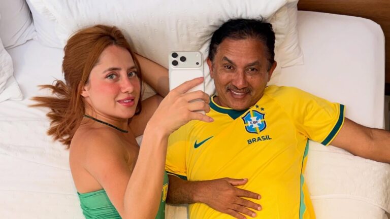 Jennefer Karoline segura celular para selfie ao lado de Manoel Gomes de camisa da seleção brasileira deitados em uma cama sorrindo
