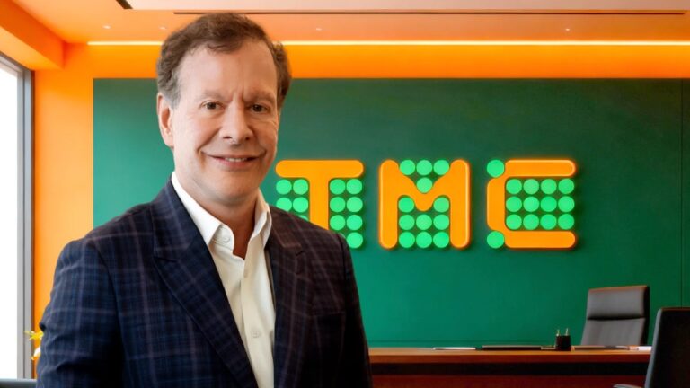 João Camargo de terno azul sorri diante de sala com logotipo da TMC ao fundo em escritório corporativo