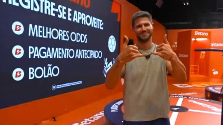 Jonas Sulzbach sorri e faz gesto com as mãos diante de painel da Betano durante ação comercial no BBB 26
