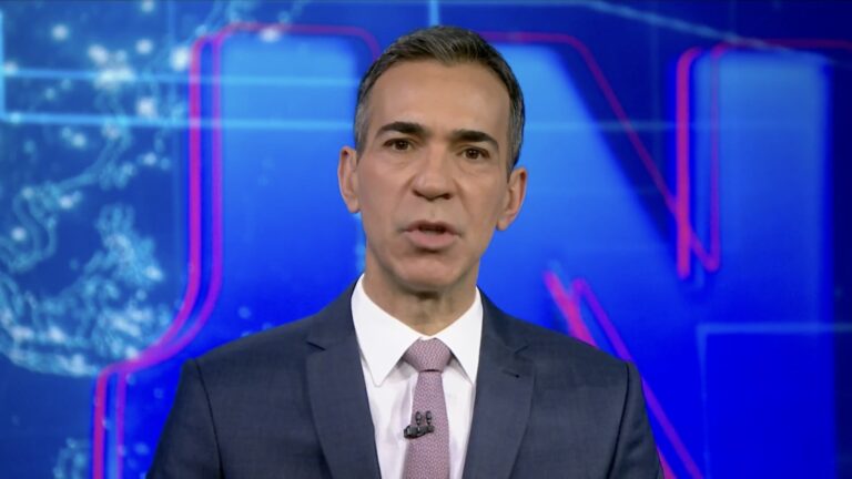 César Tralli, de terno azul e gravata rosa, fala à câmera no estúdio do Jornal Nacional com telão azul ao fundo