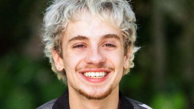 Juliano Floss sorri para a câmera com cabelo platinado e brincos diante de fundo desfocado ao ar livre