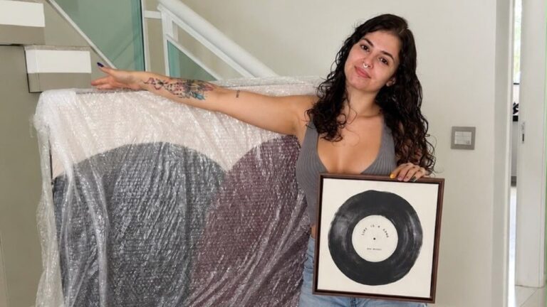 Kerolay Chaves posa de regata cinza e shorts jeans segurando quadro com disco de vinil ao lado de tela protegida por plástico em casa