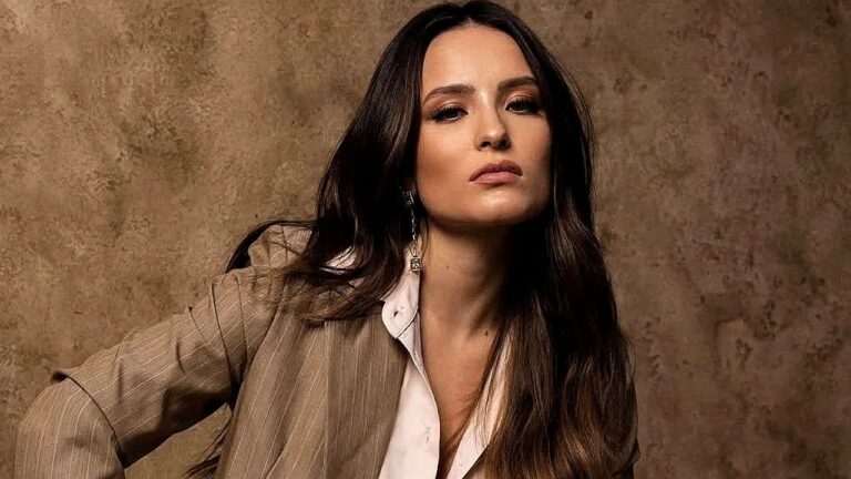 Larissa Manoela usa blazer bege e camisa branca em ensaio fotográfico com expressão séria diante de fundo marrom