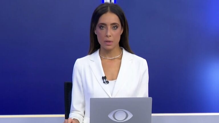 Lenny Leone de blazer branco apresenta telejornal atrás de bancada com notebook e fundo azul de estúdio