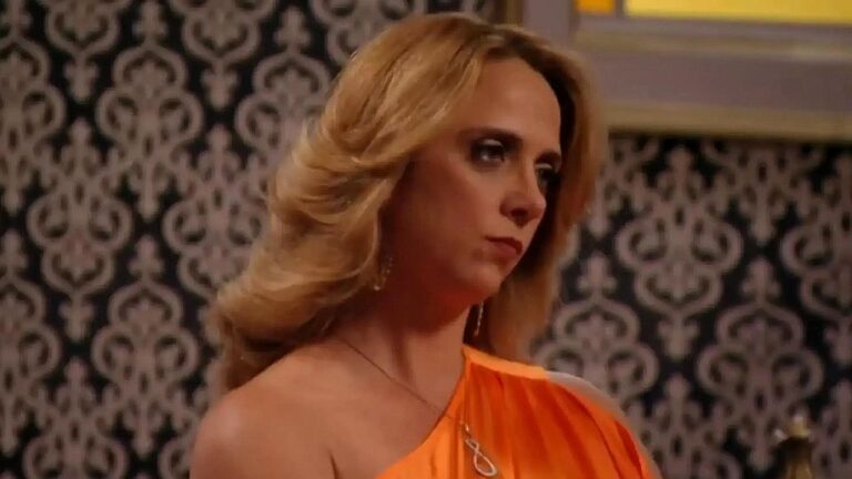Letícia Isnard com vestido laranja e expressão séria em cena de novela ambientada em sala com papel de parede clássico