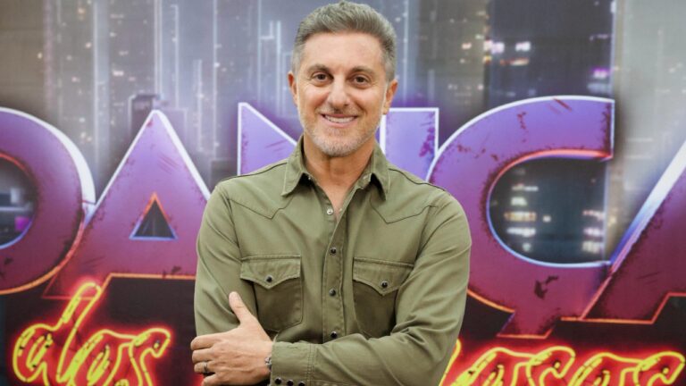 Luciano Huck de camisa verde com braços cruzados em frente a telão com logo da Dança dos Famosos