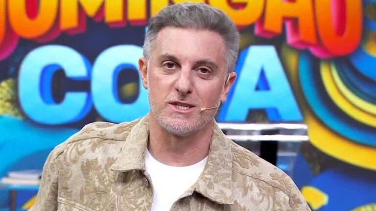 Luciano Huck de jaqueta bege e camiseta branca com microfone no rosto diante do telão do Domingão com Huck