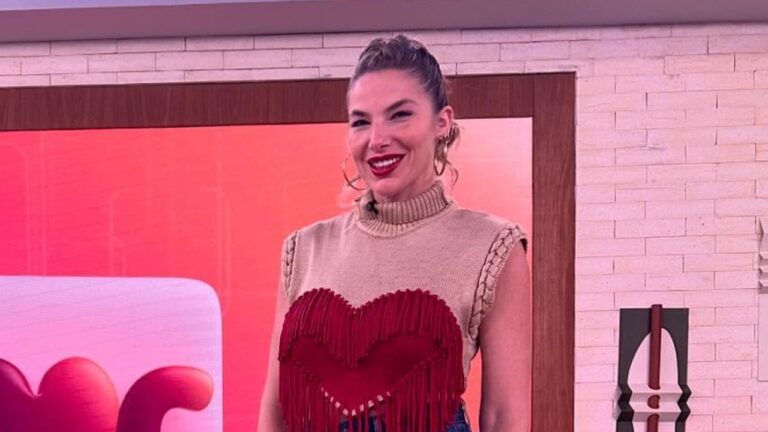 Luiza Zveiter sorri em estúdio usando blusa bege com detalhe vermelho em franjas diante de painel iluminado e parede clara