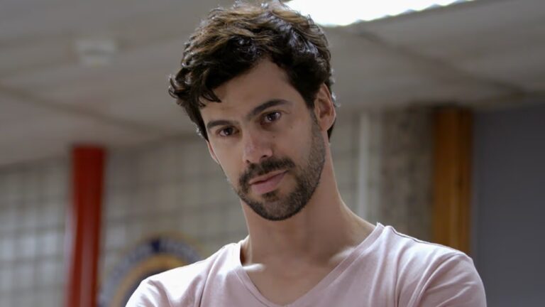 Mario De Marco de camiseta branca olha para a frente com expressão séria em ambiente interno desfocado ao fundo
