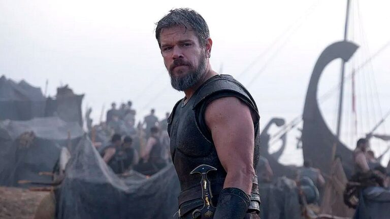 Matt Damon com armadura em cena de A Odisseia diante de embarcações e guerreiros no porto