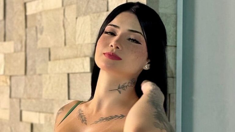 Maya Braga posa com cabelo preto solto e tatuagens visíveis no pescoço e colo diante de parede clara