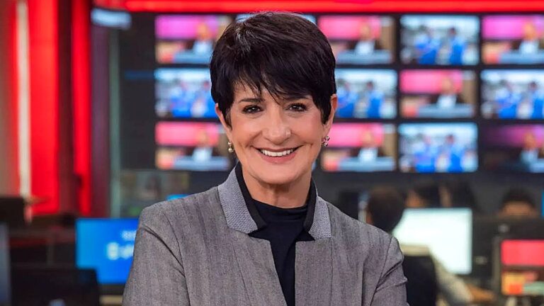 Mônica Waldvogel sorri na redação de telejornal com monitores ao fundo em imagem da jornalista
