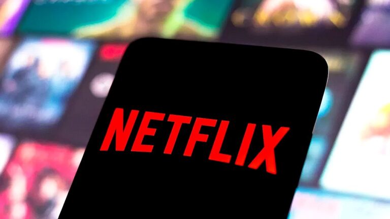 Logotipo da Netflix exibido na tela de um celular com catálogo de filmes e séries desfocado ao fundo