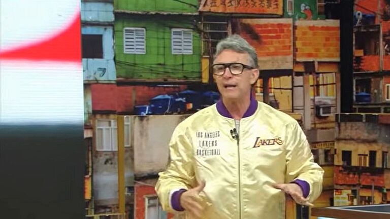 Apresentador Neto comenta no Os Donos da Bola diante de telão com imagem de comunidade ao fundo