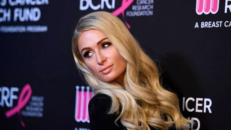 Paris Hilton posa com vestido preto e cabelos loiros em evento beneficente diante de painel de patrocinadores
